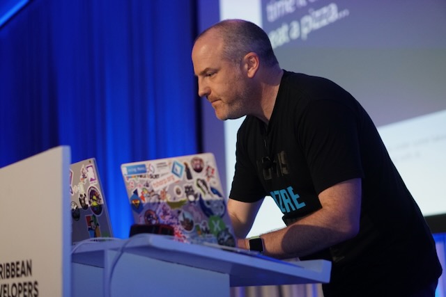A picture of Jim on a stage at a conference standing next to a podium with a laptop on it. Jim is wearing a black t-shirt and is working on the laptop