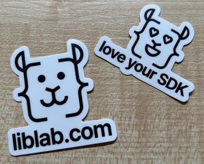 2 liblab stickers on a wooden table. One has the liblab logo, a simple line drawn llama face with curly braces for the side of the head, and liblab.com, the other has a version of the liblab llama logo with hearts for eyes and the caption love your SDK