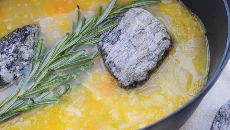 A bowl of yellow soup with a sprig of rosemary and a stone in it