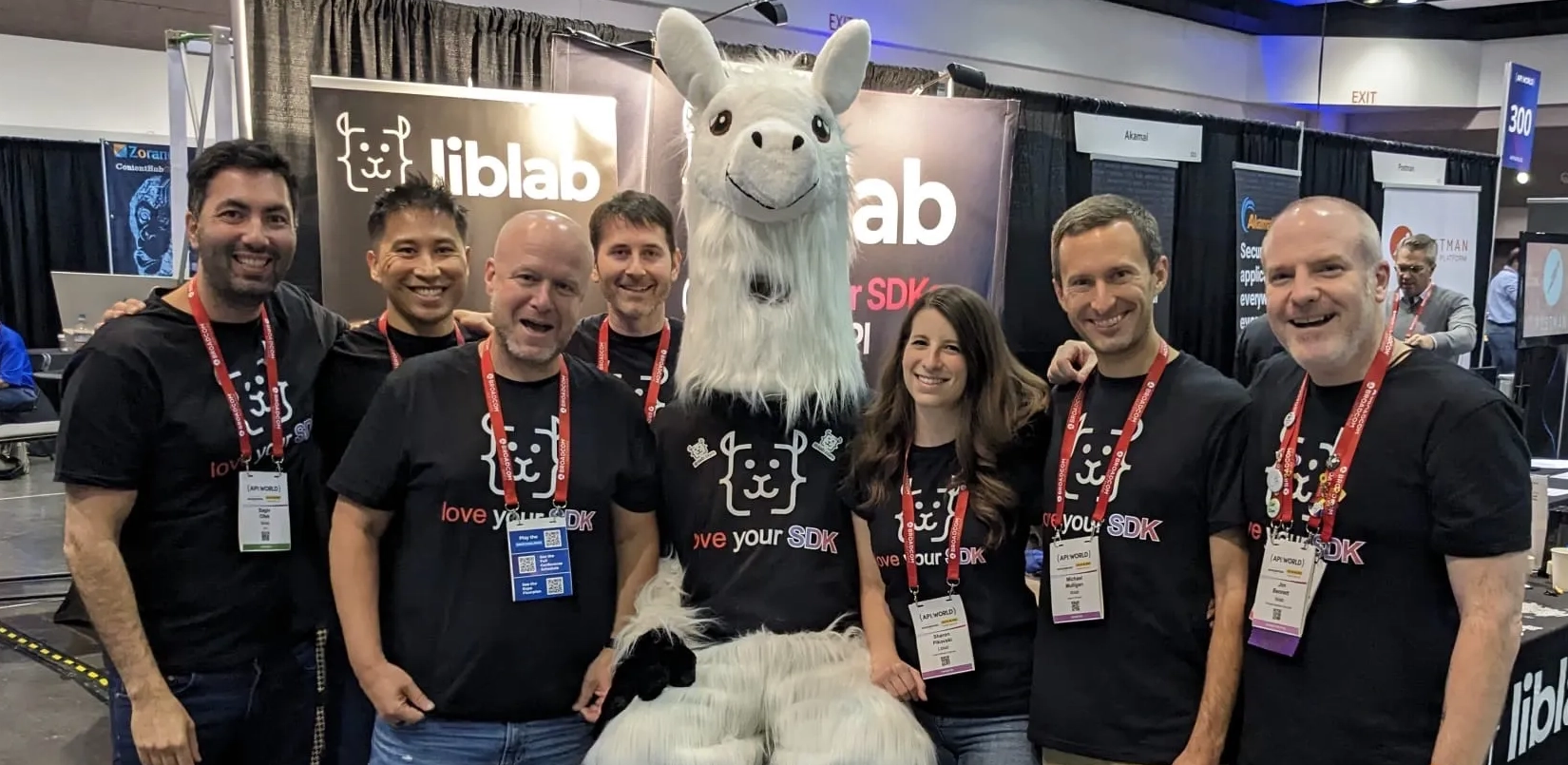 A group of people wearing liblab love your SDK shirts posing with a large llama mascot also wearing the same shirt. Behind the group is the liblab booth from API world